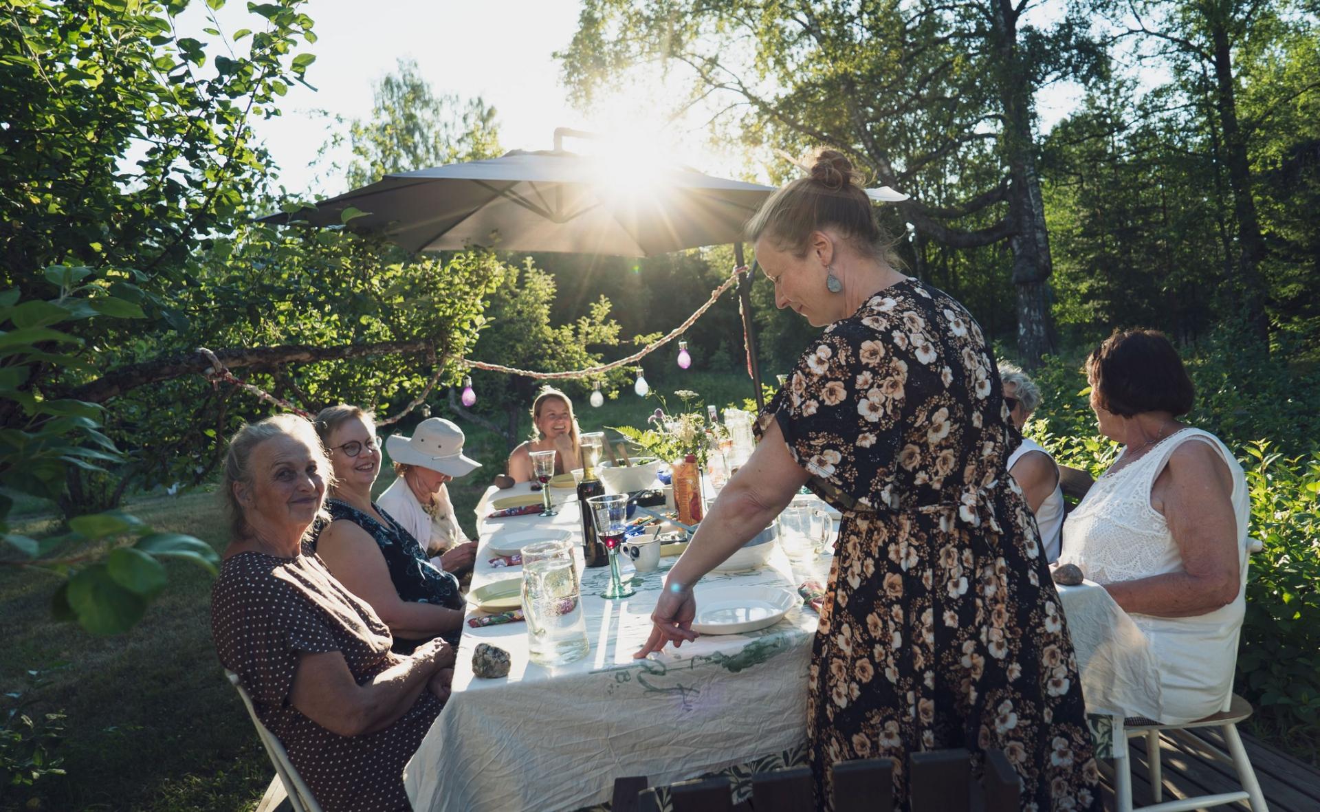 Naisia ruokapöydän ääressä puutarhassa ilta-auringon paisteessa. 
