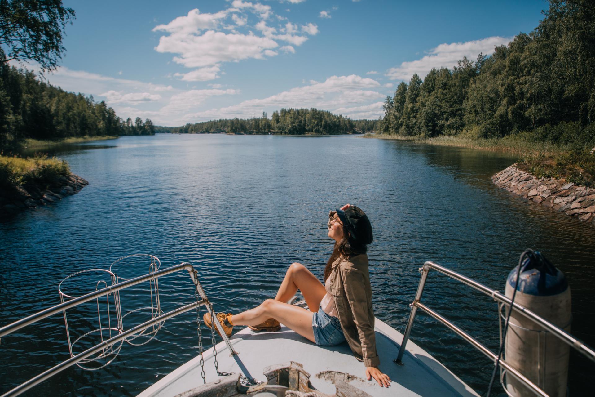 Nainen istuu veneen keulassa ja ihailee maisemia auringon paisteessa. 