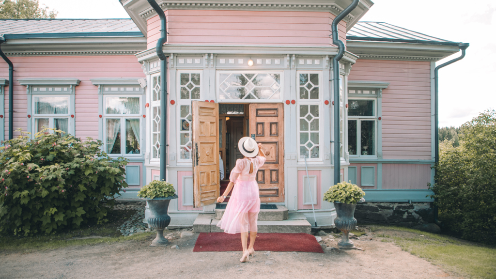 Vaaleanpunainen kartanorakennus, jonka koristeellisten verannan ikkunoiden ja avoimen edessä seisoo nainen vaaleanpunaisessa mekossa. 