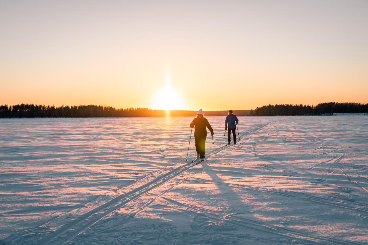 Nainen ja mies hiihtävät järvenjääladulla ilta-auringon paisteessa. 
