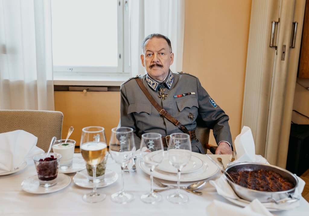 Näyttelijä esittämä Mannerheim istuu pöydän ääressä, jolle on katettu lautaset, viinilasit sekä uuniruokaa lisukkeineen. 