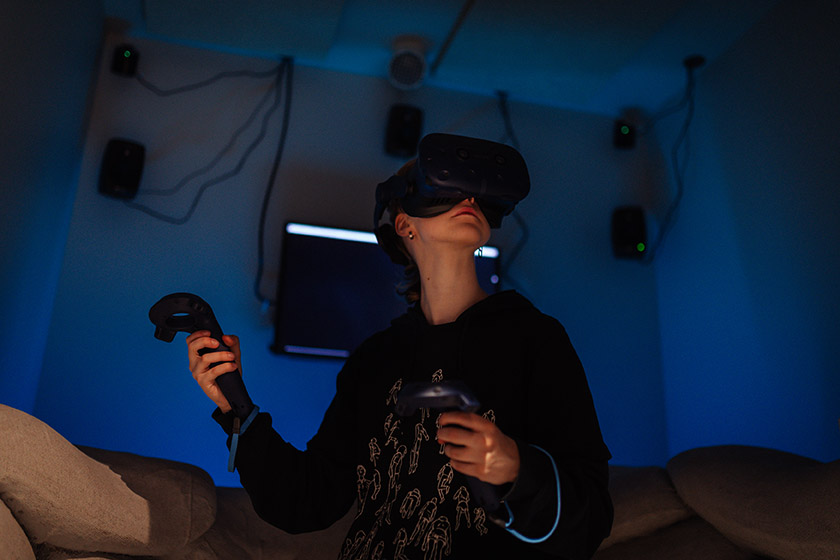 Nainen seisoo pimeässä kopissa virtuaalilasit päässä ja pitelee käsissään virtuaaliohjaimia. 
