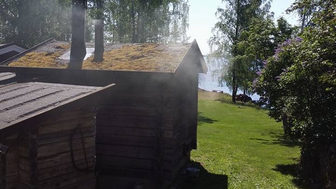A small wooden smoke sauna building is being heated by a lake.