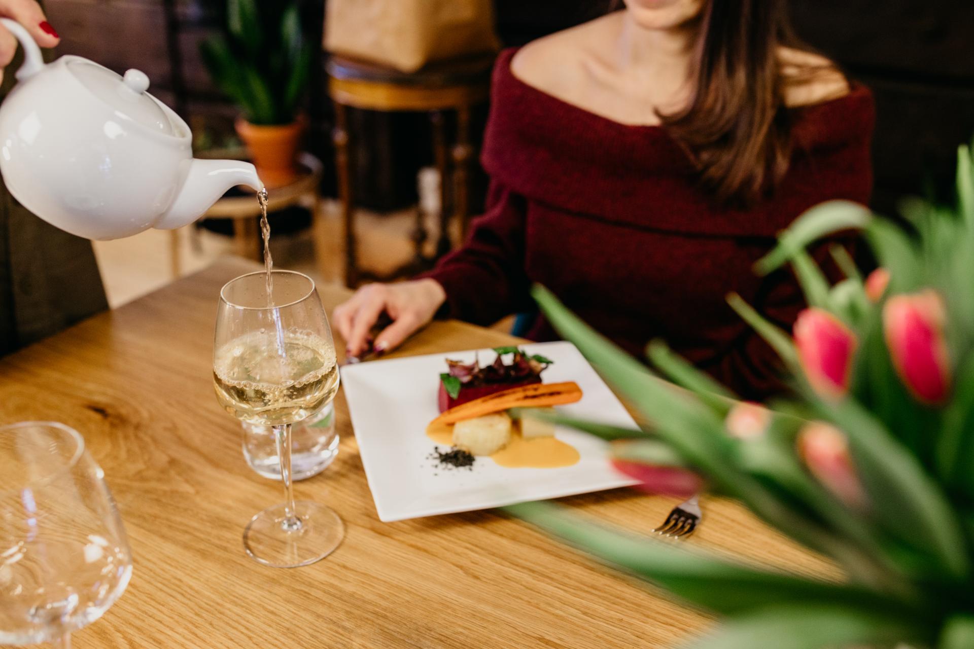 Nainen istuu ruokapöydän ääressä edessään ruoka-annos ja viinilasi, johon kaadetaan teetä. 