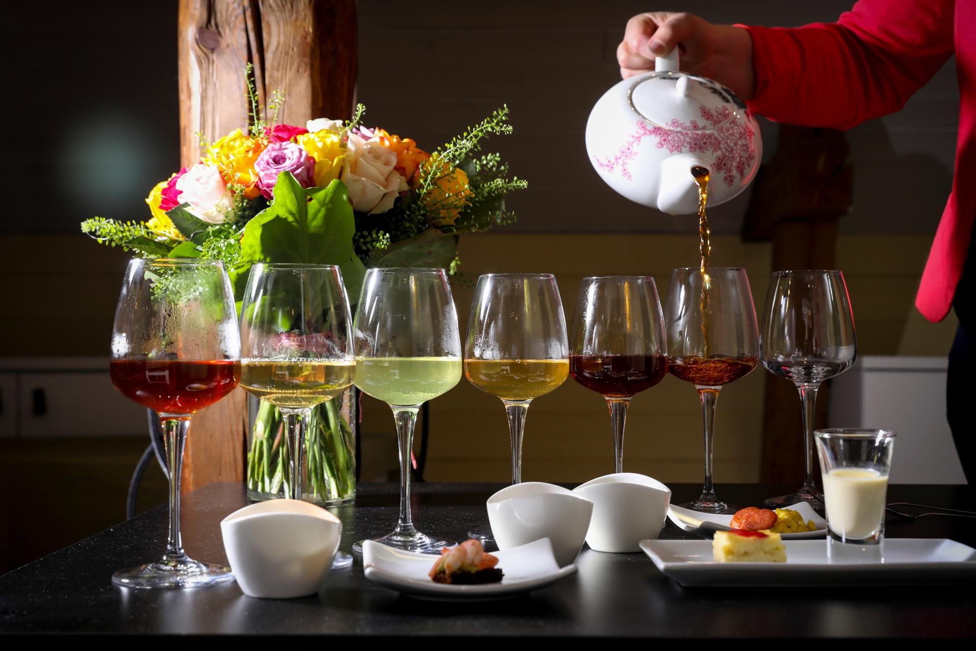 Pöydällä on kukkakimppu, pieniä suolaisia annoksia sekä viinilaseja, joissa on erilaisia teejuomia. 