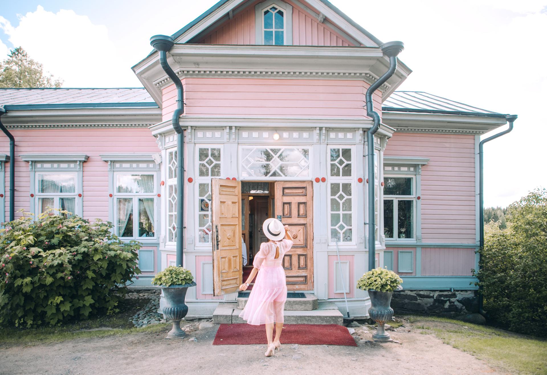 An old, wooden manor house and a woman in a pink dress standing in front of it.
