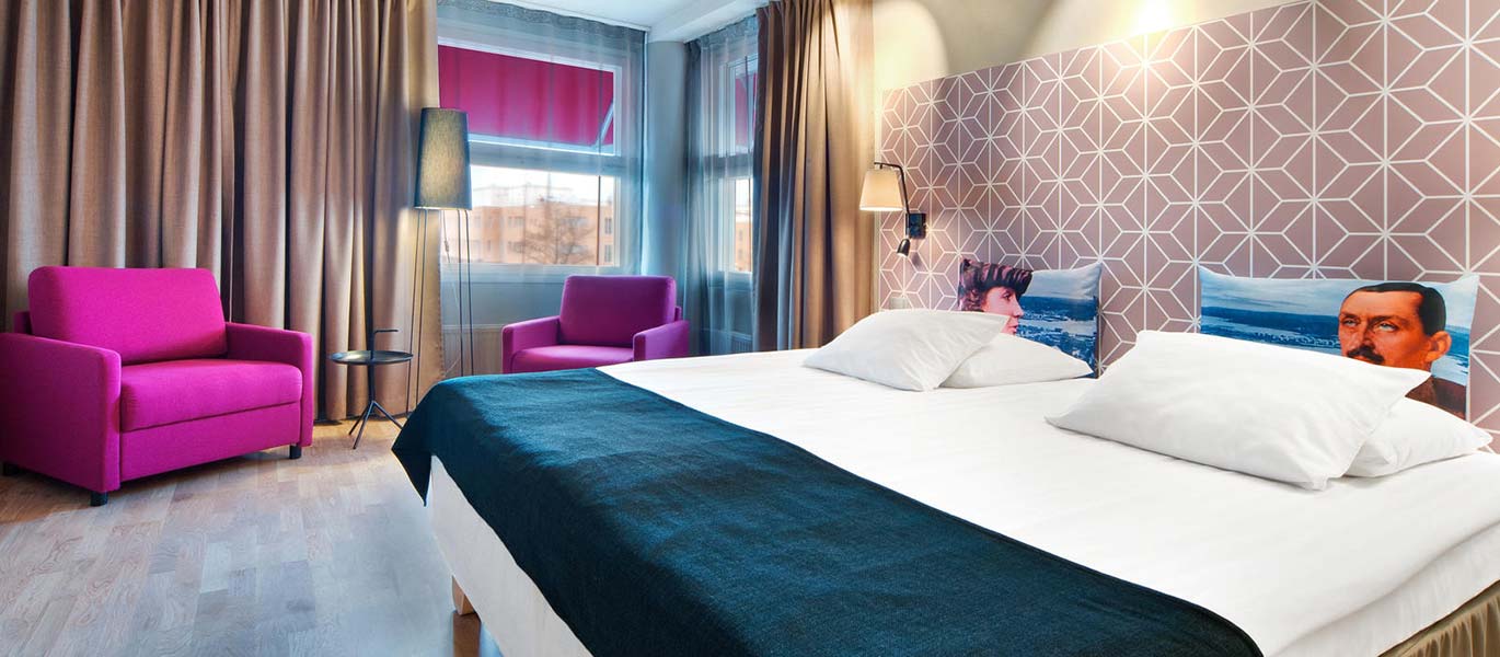 Viihtyisä hotellihuone, jossa parivuode sekä kaksi nojatuolia.