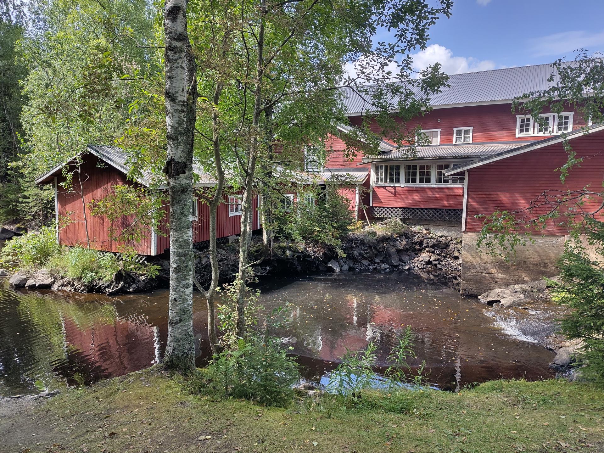 Punainen rakennuskokonaisuus hiljaa ohitse virtaavan joen takana