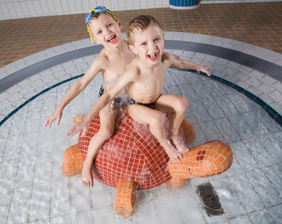 Kaksi pientä poikaa istuvat kilpikonnan muotoisen patsaan päällä pienessä altaassa ja nauravat.  